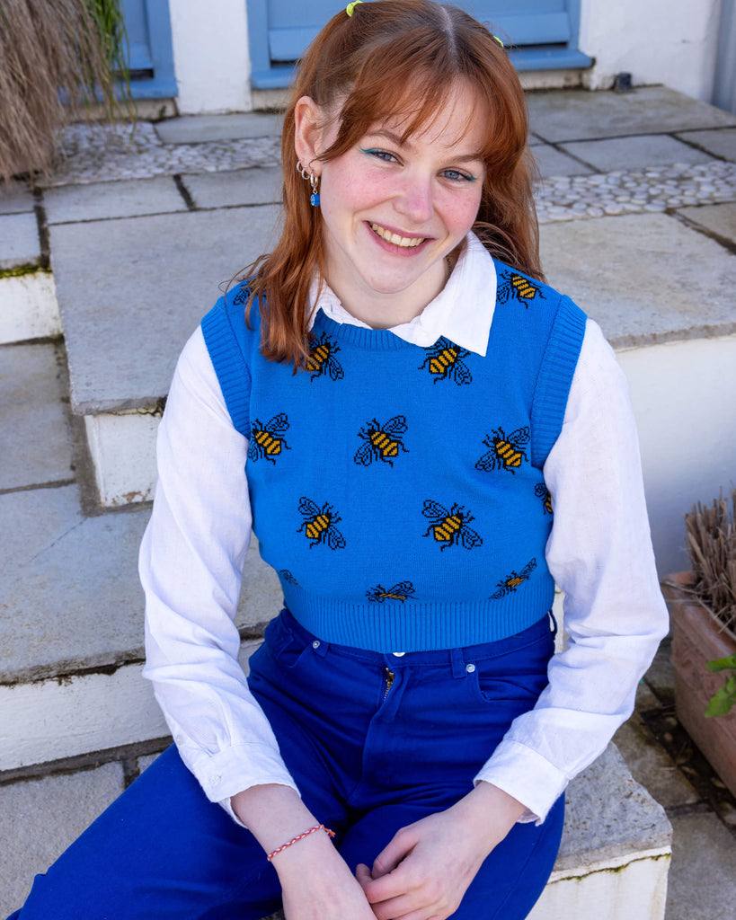 Tasha a femme red head with short hair and a fringe is smiling at camera wearing a blue cropped bee knitted top with a white shirt and bright blue jeans. She is sat by a blue door