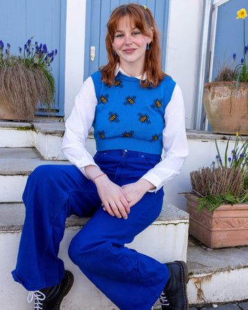 Tasha a femme red head with short hair and a fringe is smiling at camera wearing a blue cropped bee knitted top with a white shirt and bright blue jeans. She is sat by a blue door
