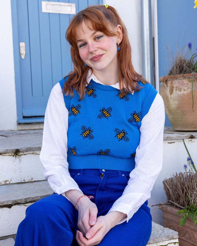 Tasha a femme red head with short hair and a fringe is smiling at camera wearing a blue cropped bee knitted top with a white shirt and bright blue jeans. She is sat by a blue door