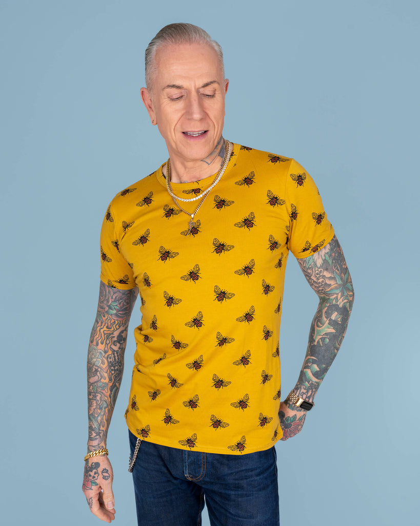 James smiling brightly, wearing a yellow bee-print T-shirt, showing the vibrant design across the chest and arms, paired with dark jeans, set against a light blue backdrop.