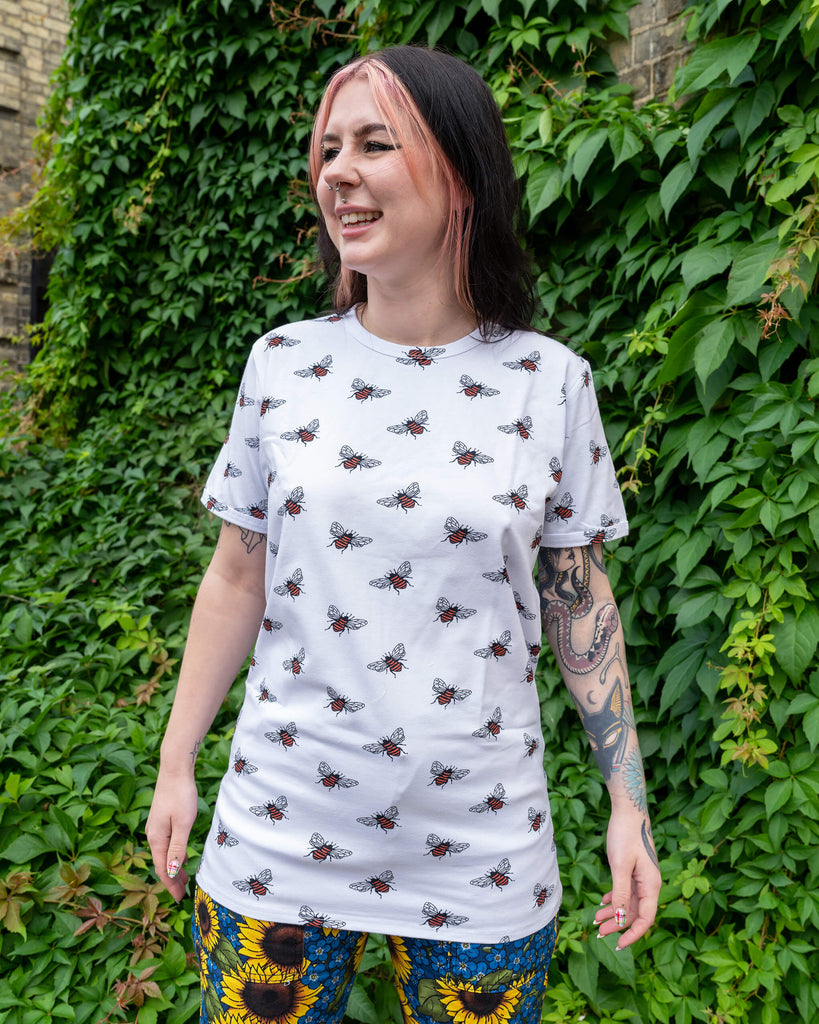 Florence is stood outside amongst greenery wearing Run & Fly White Bee Short Sleeve T Shirt paired with Run & Fly x Jen James Design Forget Me Not Charity High Waist Straight Leg Jeans. The white t shirt has an all over bumble bee print. She is smiling at the camera with her hands out by her sides

