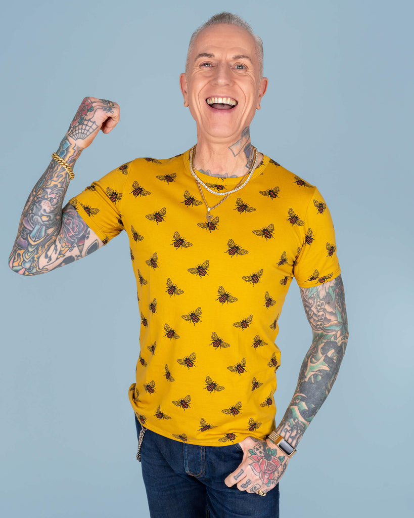 Close-up of James wearing a bright mustard yellow T-shirt with a repeat print of black and orange bees, styled with gold jewellery and visible tattooed arms, against a light blue background.