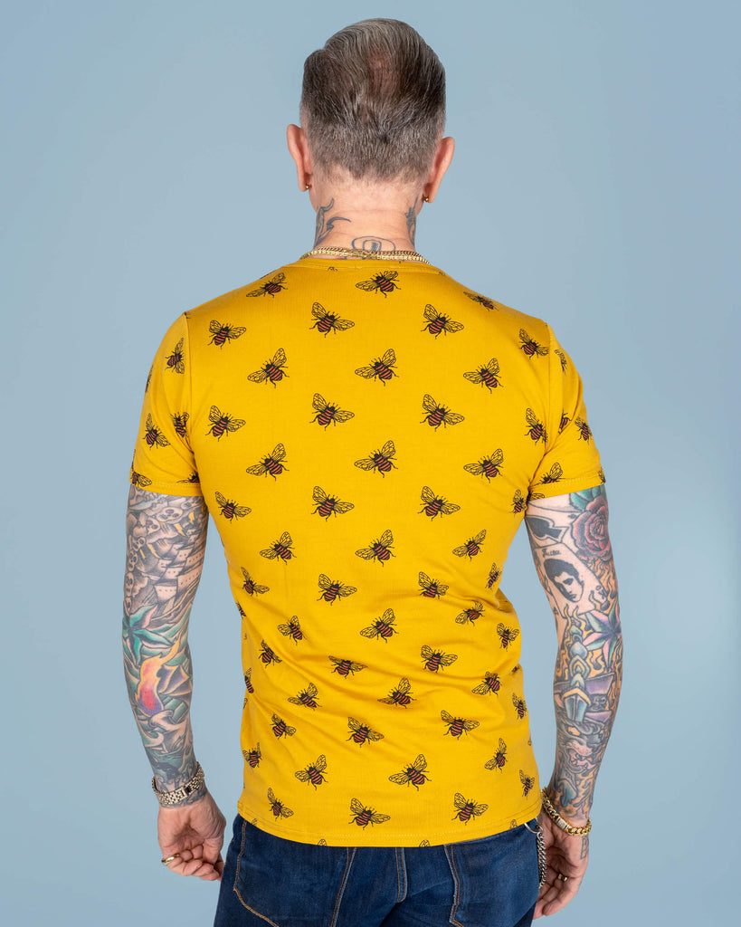 Rear view of James wearing the yellow bee-print T-shirt, showcasing the back pattern, his tattooed arms, and a neatly styled short haircut, all set against a soft blue background.