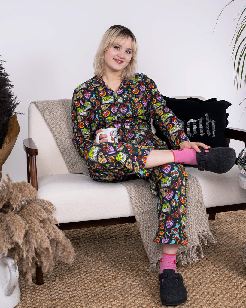 A relaxed scene with the model lounging on a sofa in the Halloween: Bewitched Stretch Twill Boiler Suit by Run & Fly. The slow fashion boiler suit features whimsical, spooky motifs and is paired with pink socks and dark slippers, creating a cosy, autumnal aesthetic in an indoor setting.