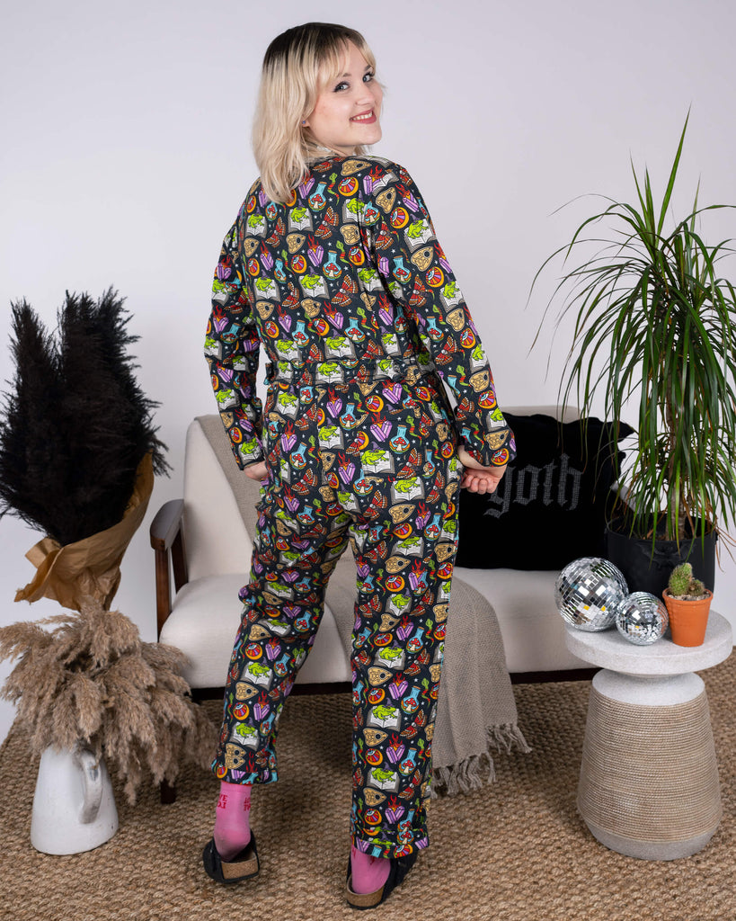 Back view of the Halloween: Bewitched Stretch Twill Boiler Suit by Run & Fly, modelled by a smiling individual. The slow fashion piece highlights playful, bold prints of magical items, perfect for autumnal style. The outfit is paired with pink socks and black shoes, framed by a cosy indoor setting.