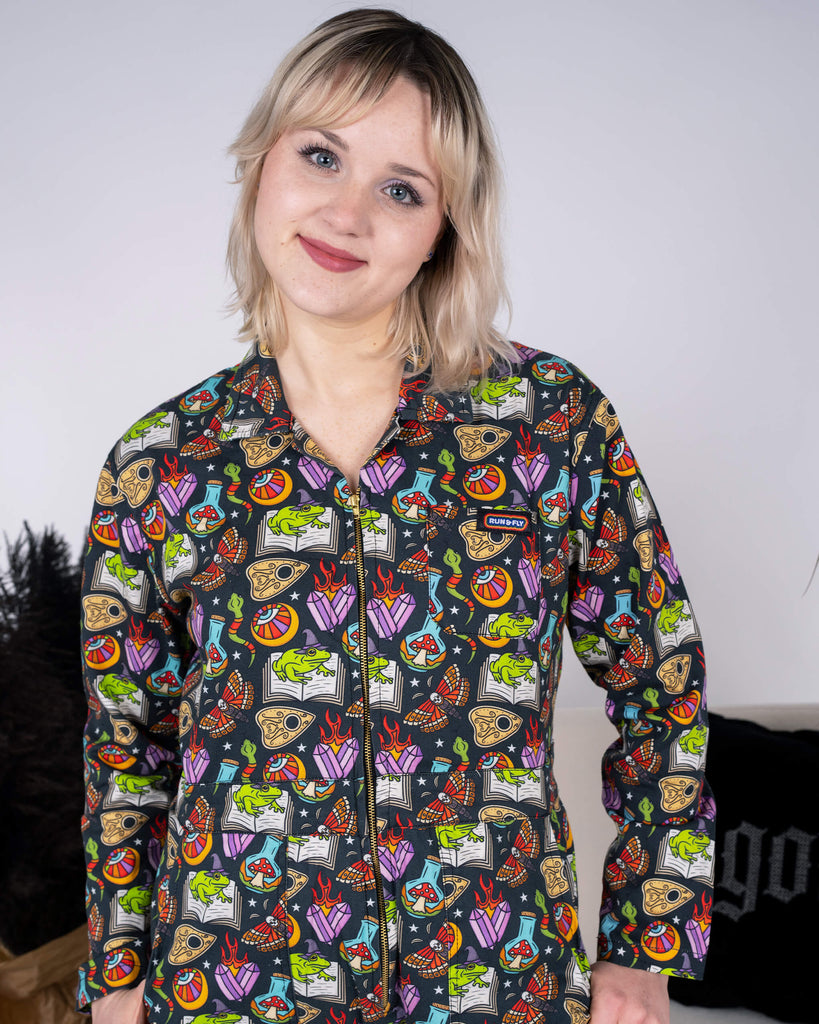 A model wearing the Halloween: Bewitched Stretch Twill Boiler Suit by Run & Fly. The ethically made jumpsuit features vibrant spooky illustrations, including frogs, books, and flames, on a black background. The model stands against a minimalist backdrop, showcasing the zip-front design and long sleeves of the comfortable, sustainable outfit.