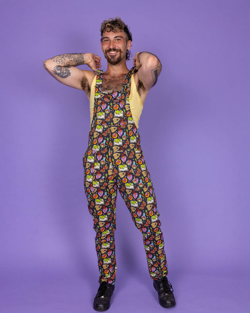 Masc non binary model wearing Run & Fly 'Bewitched' print dungarees in a joyful pose with arms behind the head. The vibrant all-over design features witchy motifs, including cauldrons, spell books, and potion bottles. Ethical and quirky, Run & Fly is a small slow fashion company known for their unique prints and eco-friendly clothing.