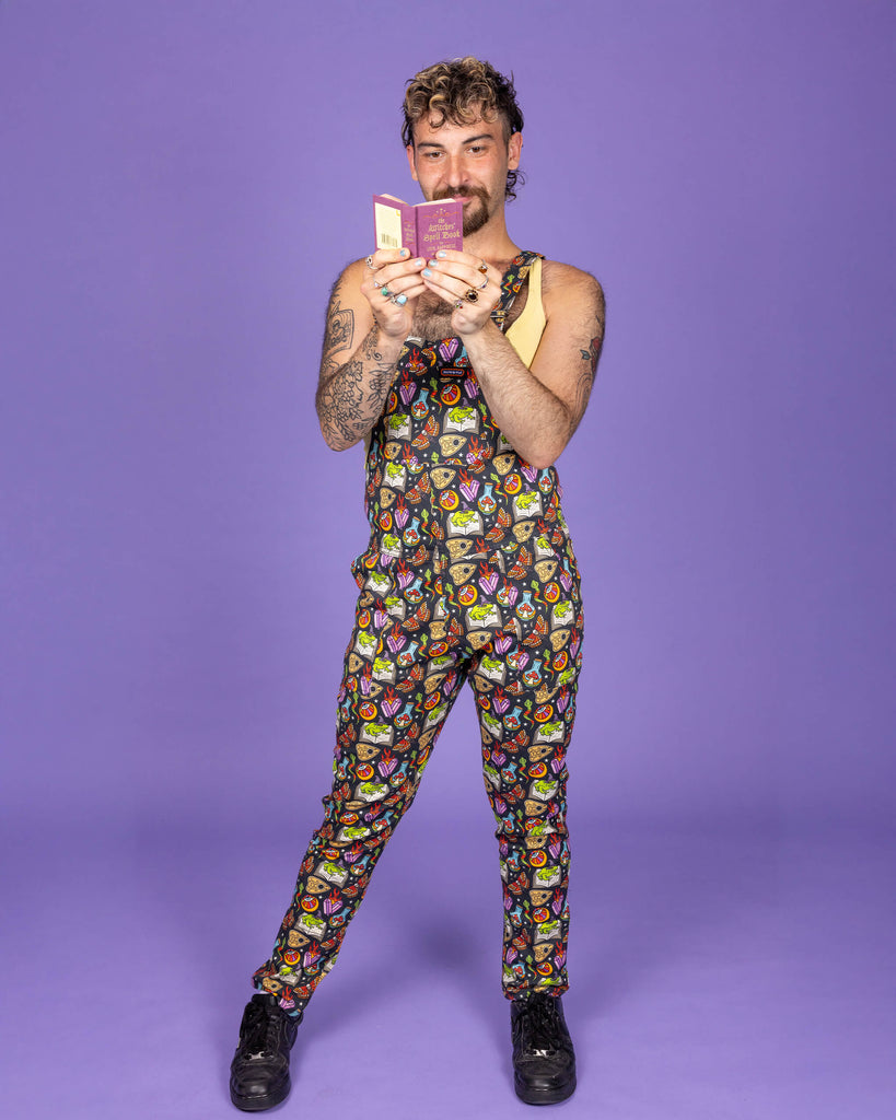Masc non binary model playfully holding a mini spell book while wearing Run & Fly's 'Bewitched' dungarees. The all-over print features magical elements like cauldrons, crystals, and spell books. Run & Fly is a small ethical, slow fashion brand known for their quirky and fun designs.