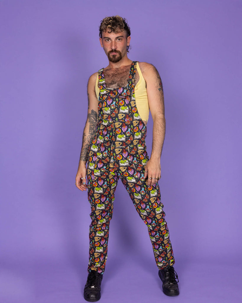 Full body shot of a masc non binary model confidently standing in Run & Fly's 'Bewitched' print dungarees, featuring a witchy-themed all-over design with cauldrons, potion bottles, and skulls. The model pairs it with a yellow top, highlighting the fun, playful energy of Run & Fly's ethical slow fashion approach.