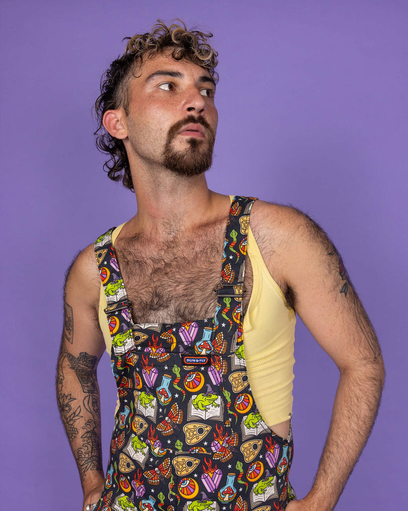 A masc non binary model wearing Run & Fly 'Bewitched' dungarees featuring vibrant witchcraft imagery. The chest pocket and straps display a print filled with mystical elements like spell books, cauldrons, and magical crystals. The ethical, small business Run & Fly stands out with quirky, slow fashion designs.
