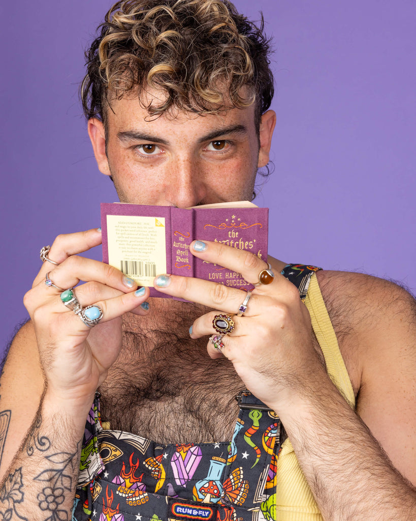Close-up of a masc non binary model with a brown curly mullet wearing Run & Fly 'Bewitched' dungarees while holding a mini spell book. The colourful print features witchcraft-inspired imagery like skulls, potion bottles, and magic symbols. Run & Fly creates fun and playful fashion, supporting ethical, slow fashion practices.