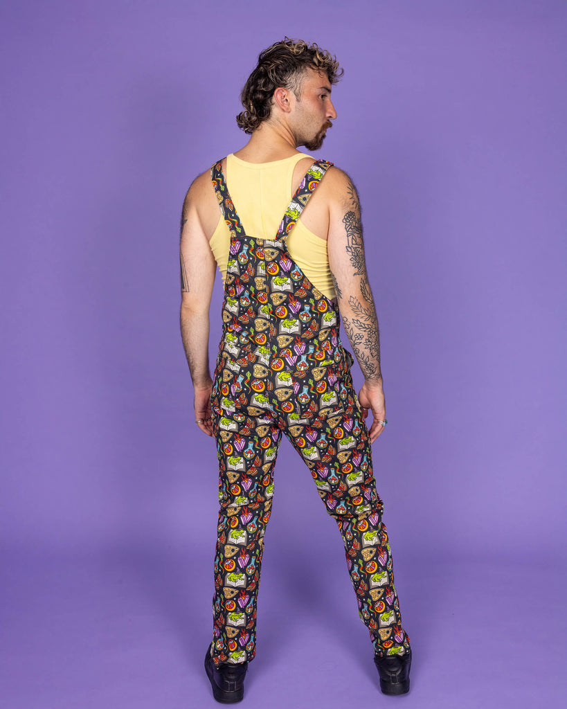 Masc non binary model with brown curly mullet hair wearing Run & Fly 'Bewitched' print dungarees with a mystical, witchy-themed design, featuring cauldrons, spell books, and potion bottles. The model stands confidently, back turned, showcasing the adjustable straps and all-over vibrant print. Ethical, slow fashion company Run & Fly is known for their unique and quirky designs.