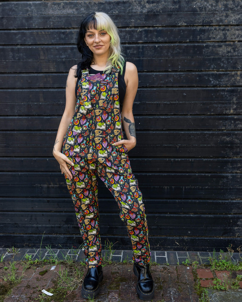 Full-body shot of a person wearing Run & Fly dungarees with a vibrant, whimsical print, including frogs, crystals, and mystical symbols. The person is standing against a dark wooden background, showcasing their alternative fashion style with platform black boots.