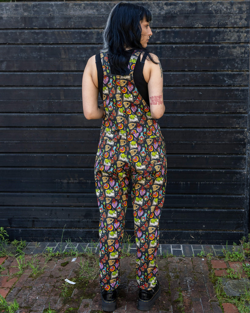 Full back view of a person wearing Run & Fly dungarees with a vibrant, magical print. The pattern includes elements like frogs on books, eyes, mushrooms, and crystals. The person has black and blonde hair, styled in waves, and is standing against a dark wooden background.