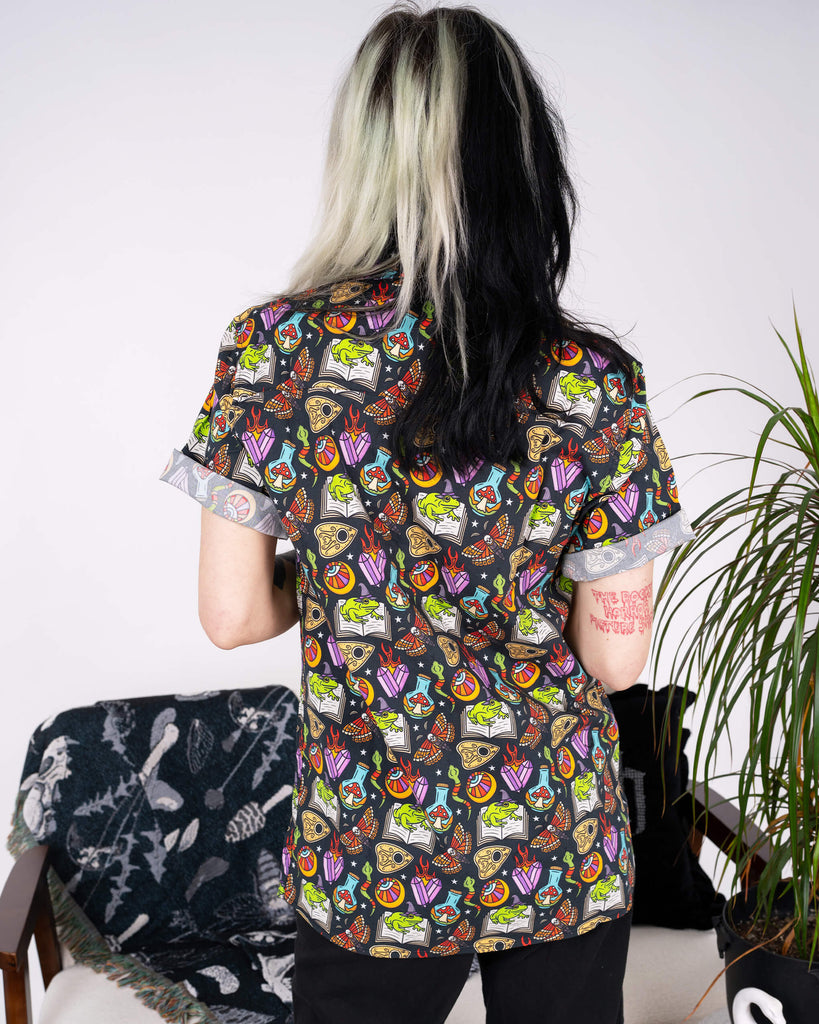 A back view of the Run & Fly Halloween: Bewitched Short Sleeve Shirt, showcasing its full design. The print includes colourful Halloween motifs like witches’ hats, bubbling cauldrons, and mystical symbols on a black base. The model’s black-and-blonde hair adds an alternative edge to the look. The eco-conscious, slow fashion company Run & Fly brings a whimsical twist to Halloween style.