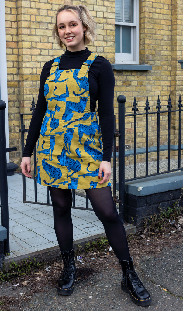 Amy is standing in front of a brick townhouse and black railings wearing the big cats stretch twill pinafore dress in yellow and blue with front pockets, paired with a black high neck long sleeve to, black tights and chunky boots. Model is facing the camera and smiling with hands by their sides.