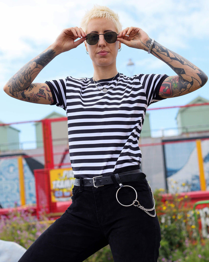 Close-up of YO wearing a black-and-white striped T-shirt, layered necklaces, and edgy accessories. Their tattoos and sunglasses add a rebellious touch to the bright, casual setting.