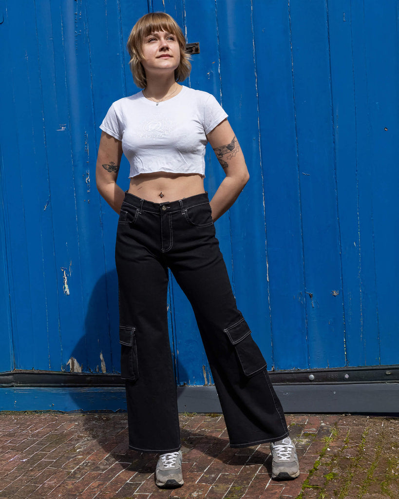 The Black Contrast Stitch Wide Leg Stretch Cargo Trousers modelled by a femme person with short blonde hair, a white crop top baby tee and grey trainers. She is stood posing in front of a blue wooden door outside leaning on her left leg, both hands resting behind her hips smiling looking off to the right. The black cargo trousers have white contrast stitching with two front flap pockets on the lower legs.