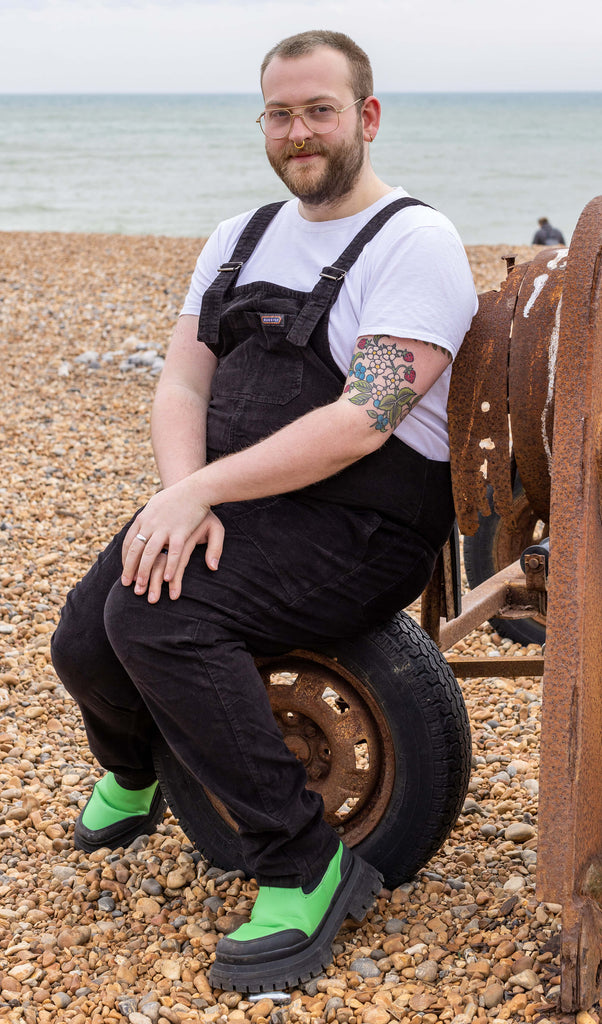 Thomas a white male with tattoos and nose ring and glasses is looking at camera smiling wearing black corduroy dungarees and green boots
