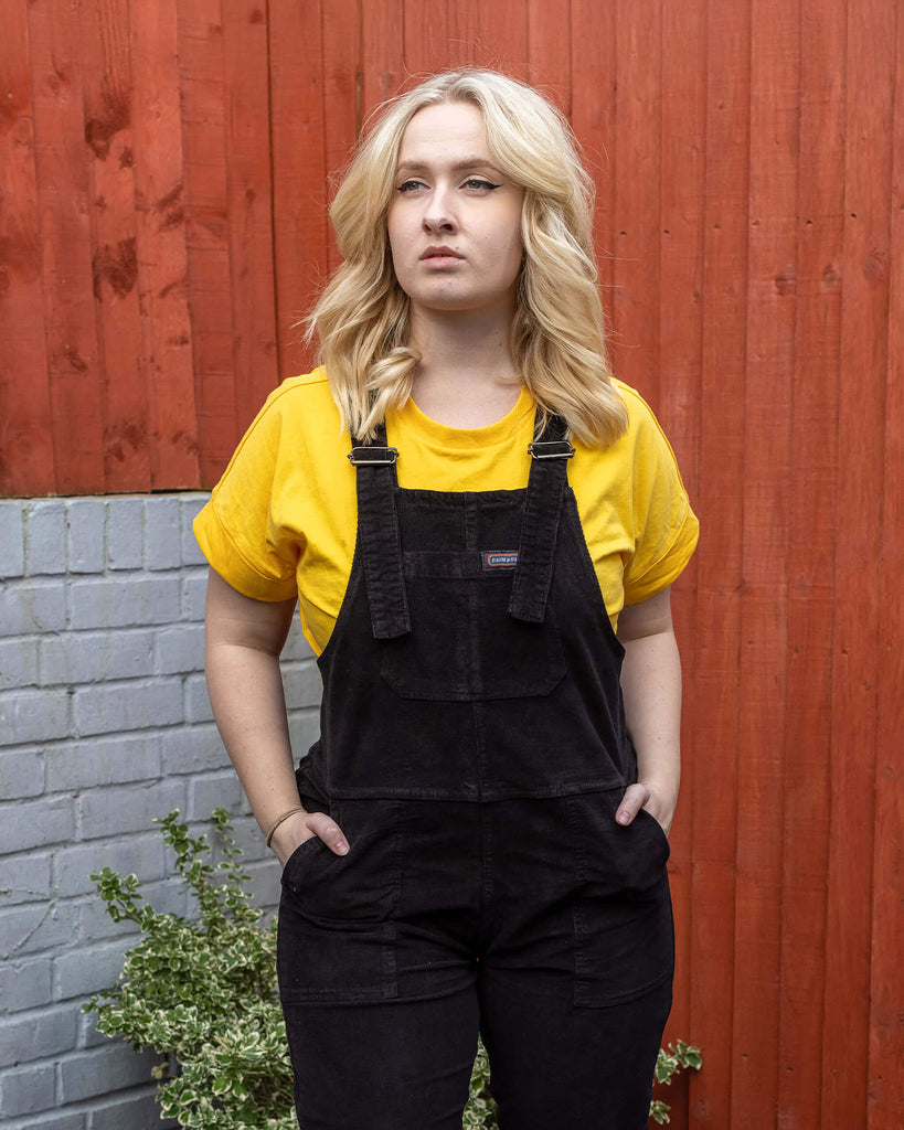 Close up of Holly a white female model with wavy blonde shoulder length hair is looking off to one side wearing black corduroy dungarees and a yellow tee underneath in a garden setting.