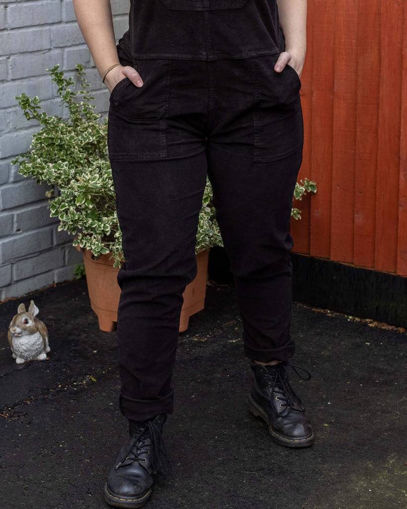 Close up of Holly a white female model facing forward with both hands in the pockets of wblack corduroy dungarees with black boots in a garden setting.