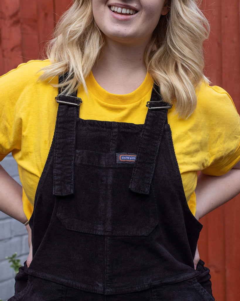 Close up of Holly a white female model with wavy blonde shoulder length hair smiling wearing black corduroy dungarees and a yellow tee underneath in a garden setting.
