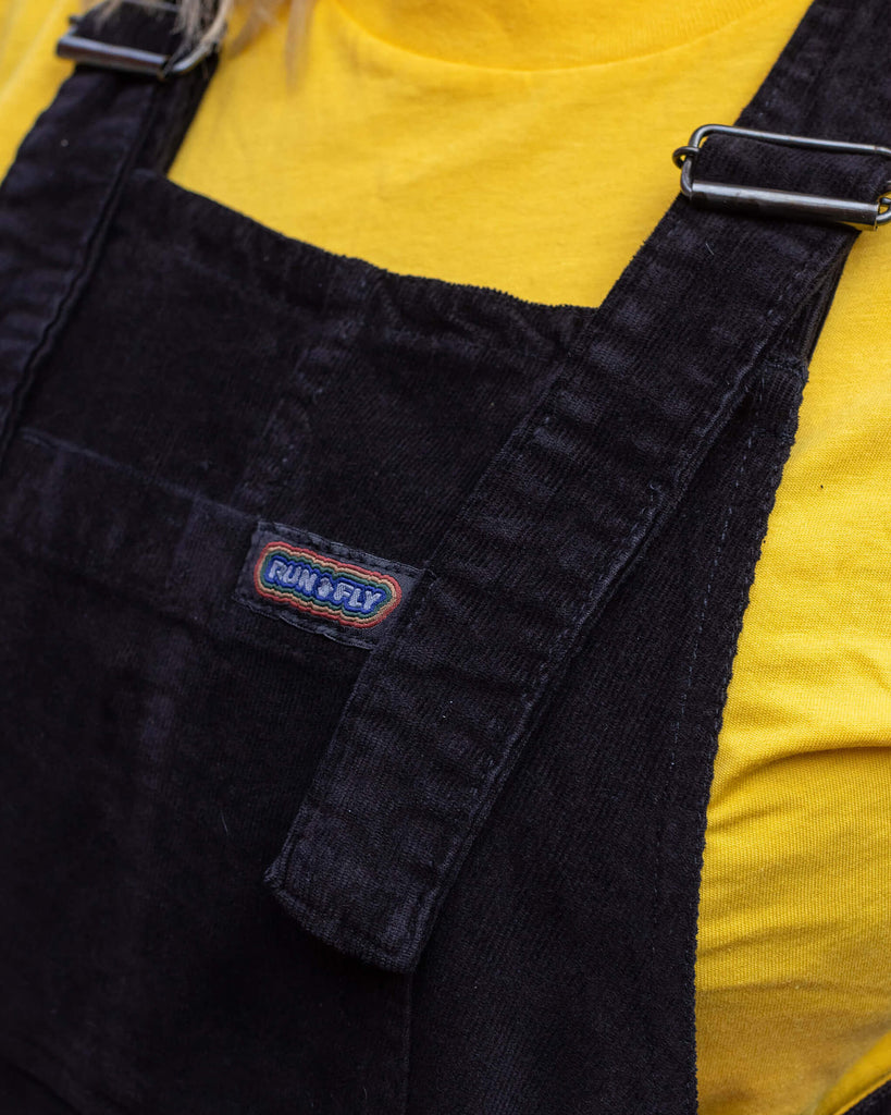 Femme model wearing black corduroy dungarees and a yellow tee underneath in a garden setting this is a close up of the Run & Fly label sewn onto the front of the dungarees.
