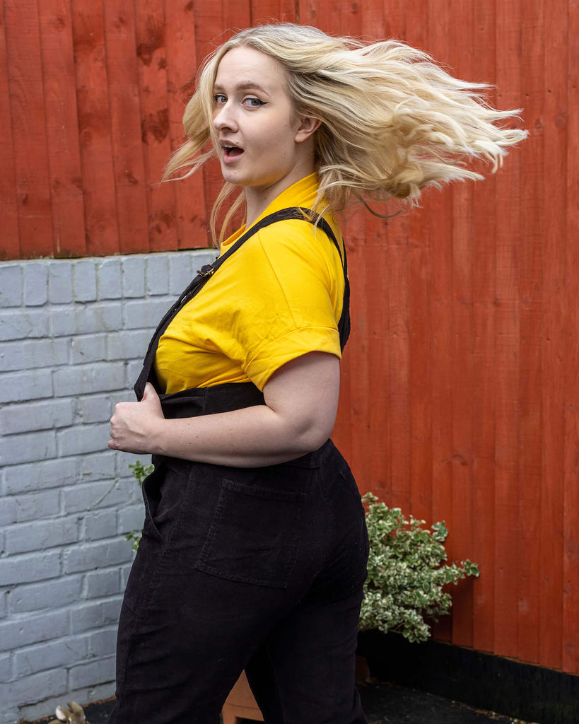 Holly a white female model with wavy blonde shoulder length hair is facing away flicking their hair over their shoulder smiling wearing black corduroy dungarees and a yellow tee underneath in a garden setting. She is whipping her hair round and looking into camera.