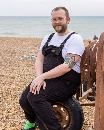 Thomas a white male with tattoos and nose ring and glasses is sat down on brighton beach looking at camera smiling wearing black corduroy dungarees and green boots.