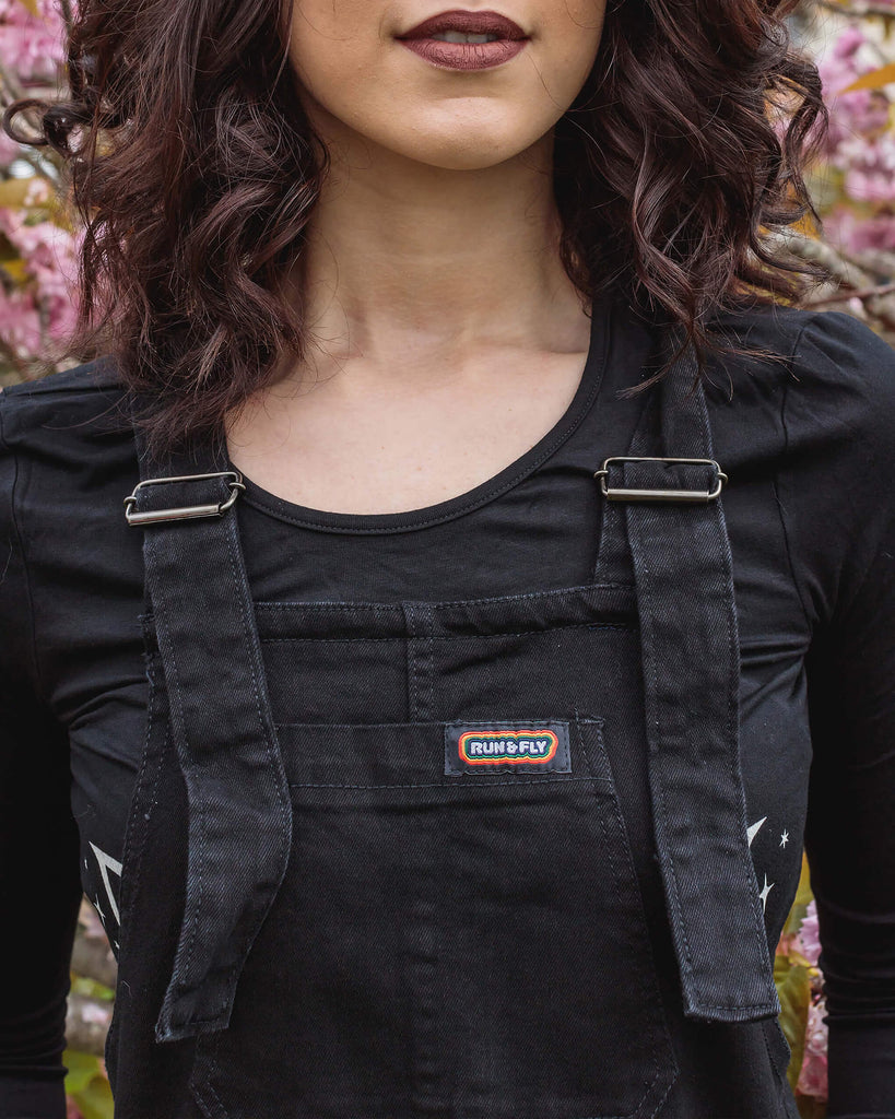 Close up of Sophie wearing the Black Stretch Denim Dungarees with a long sleeve black tee in front of a pink blossom tree.