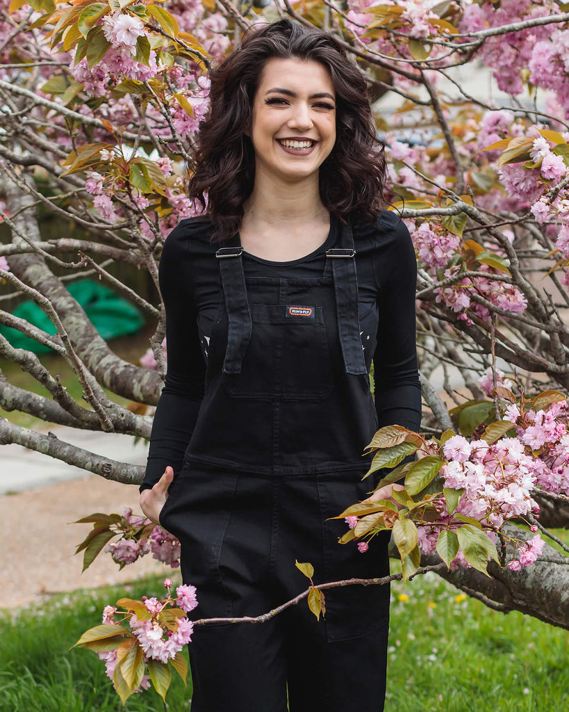 Close up of Sophie wearing the Black Stretch Denim Dungarees with a long sleeve black tee in front of a pink blossom tree.
