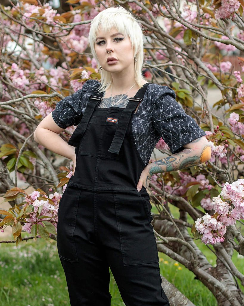 Florence with a white blonde mullet is standing with her hands on hips, in front of a pink blossom tree wearing the Black Stretch Denim Dungarees with a puff black shirt underneath and platform black boots.
