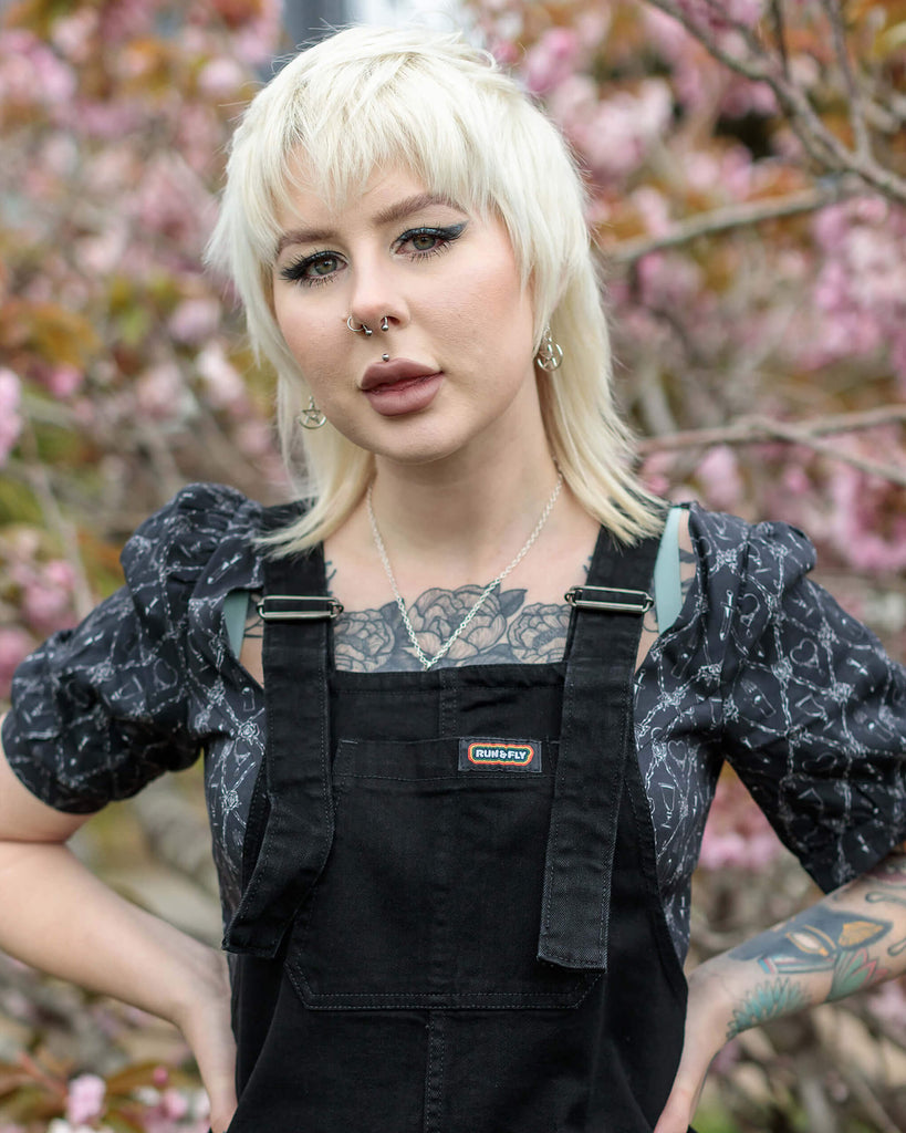 flo a blonde haired model with tattoos and a mullet is wearing black denim dungarees with a black top underneath and is standing against a pink blossom tree.