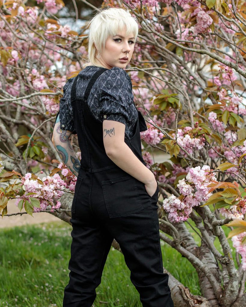 Florence with a white blonde mullet haircut is standing facing away from camera looking over her shoulder with both hands in the pockets of the Black Stretch Denim Dungarees with a puff black shirt underneath and platform boots.