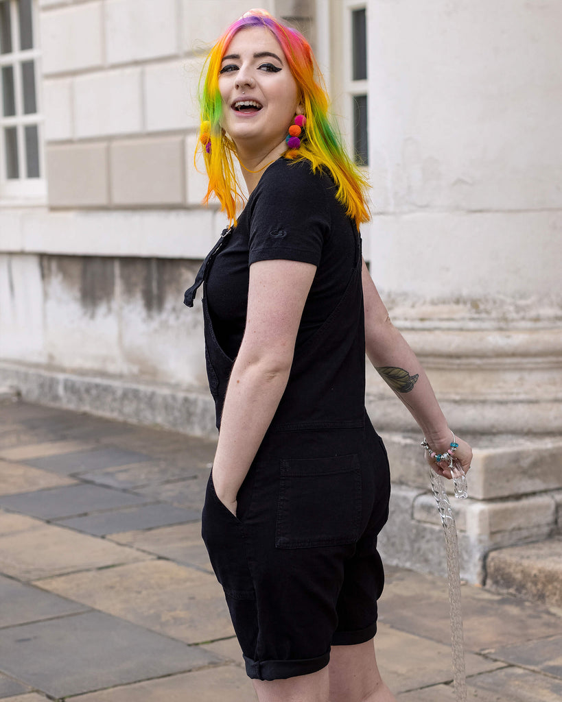 Eliza, a non binary femme person with rainbow hair using a clear sparkly walking stick, and wearing the Black Denim Stretch Dungaree Shorts with a black tee underneath and multicolour pompom earrings. They are smiling looking over their shoulder laughing outside in a cobble street in london posing facing away.