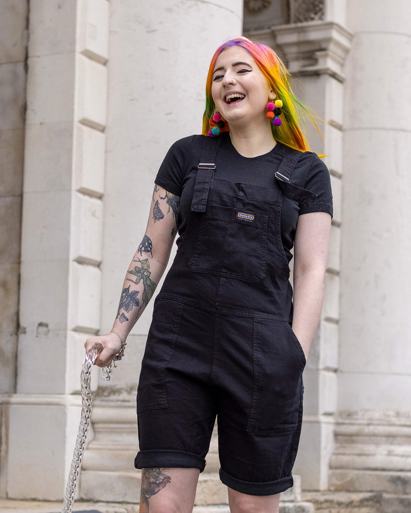 Eliza, a non binary femme person with rainbow hair using a clear sparkly walking stick, and wearing the Black Denim Stretch Dungaree Shorts with a black tee underneath and multicolour pompom earrings. They are laughing taking a step outside in a cobble street in london posing facing forward.