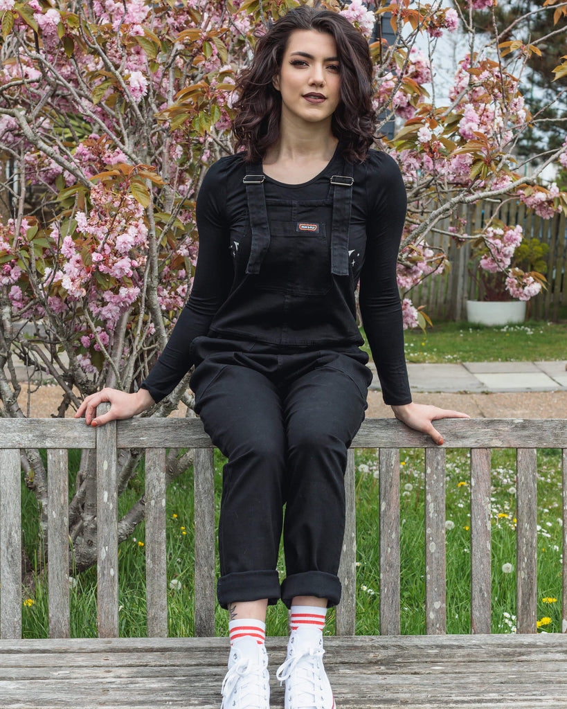 Sophie with mid length brown hair smiling sitting on a park bench by a pink blossom tree wearing the Black Stretch Denim Dungarees with a long sleeve black t-shirt and white trainers. 