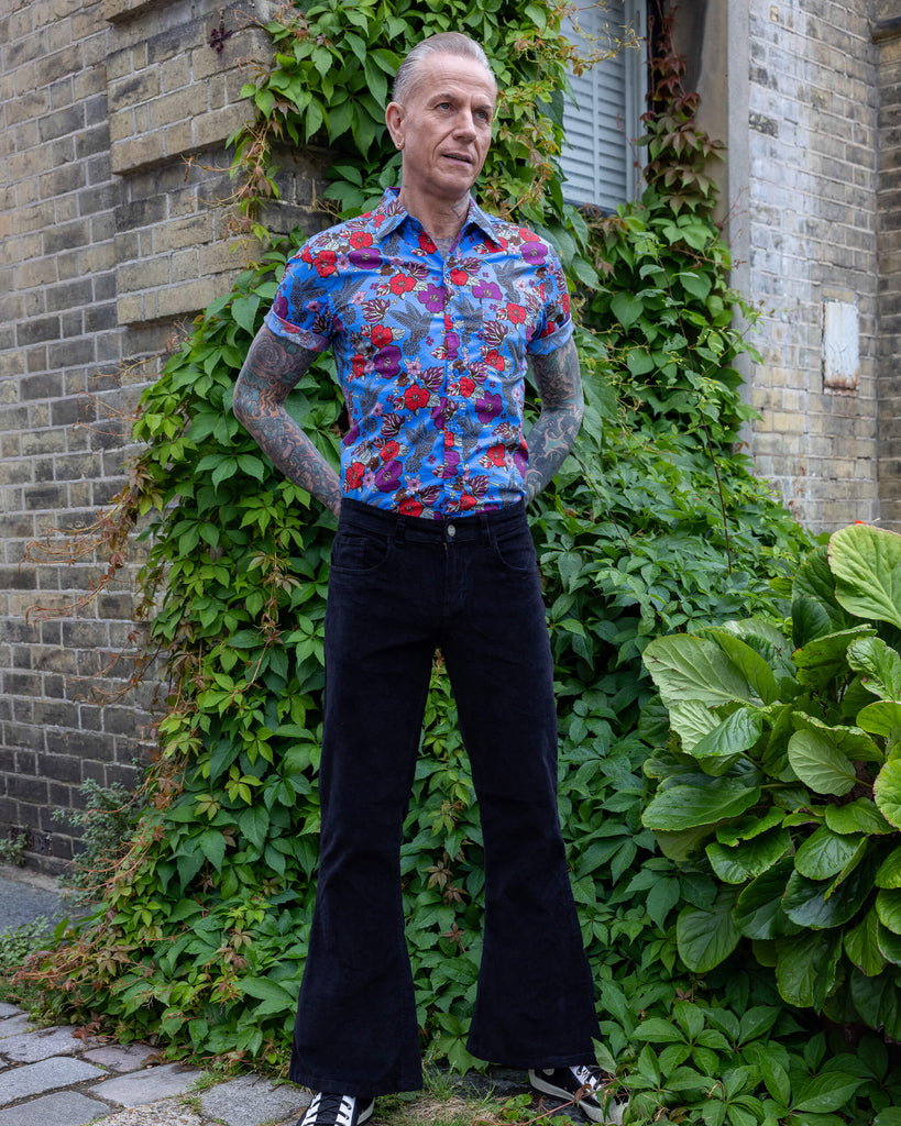 James is wearing Black Corduroy Bell Bottom Flares, paired with Hummingbird Print Short Sleeve Shirt and white trainers. He is posing with hands on his hips.