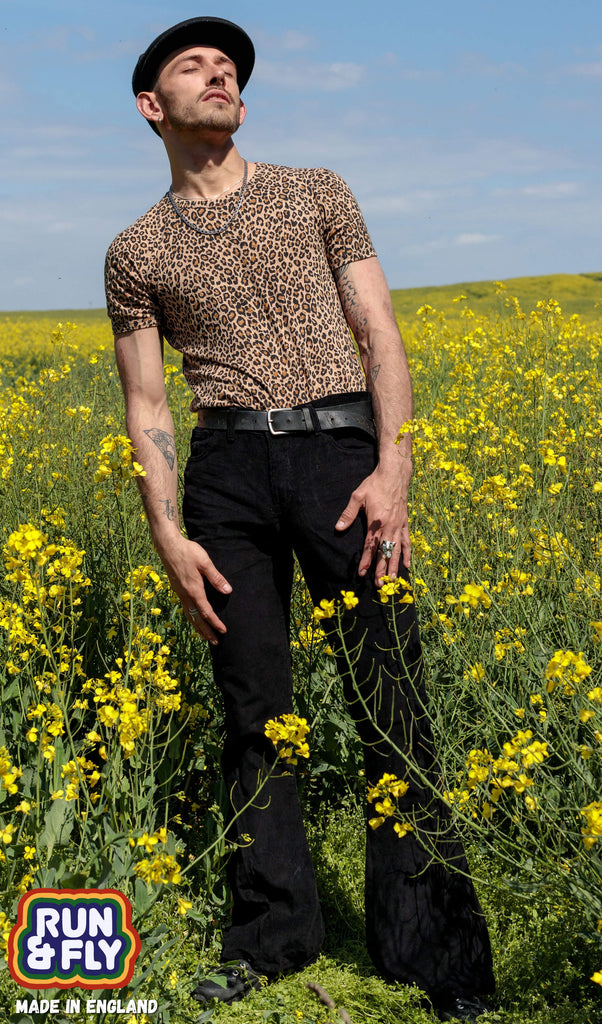 Model is stood in a yellow flower field wearing black corduroy bell bottom super flares with a natural leopard print t-shirt, black beret cap and black trainers. They are facing the camera and looking off to the right with their eyes closed.