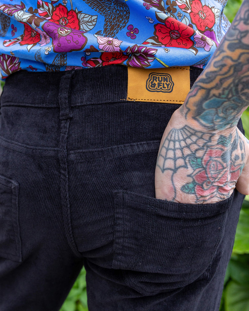 Close up of James wearing Black Corduroy Bell Bottom Flares, paired with Hummingbird Print Short Sleeve Shirt and white trainers. Close up shows the Run & Fly label on the back of the flares and model's tattooed hand in the back pocket