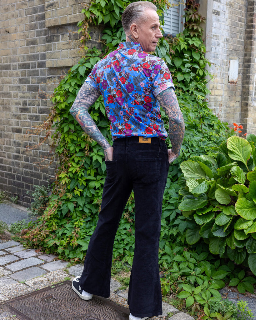 James is wearing Black Corduroy Bell Bottom Flares, paired with Hummingbird Print Short Sleeve Shirt and white trainers. He is posing with hands on his hips and looking over his shoulder.