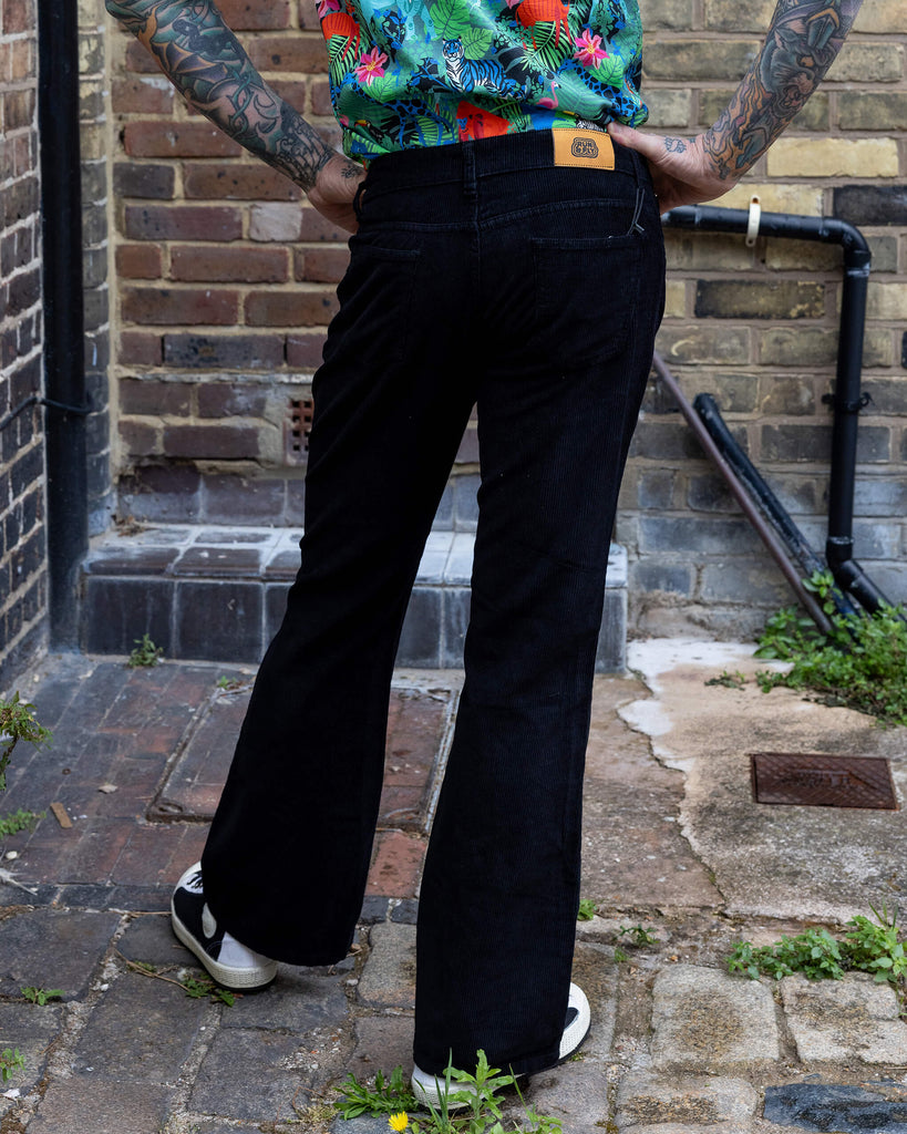 Jim is standing in a mews in Hove he is a tattooed grey haired man he is wearing a jungle print shirt and black flares by Run & Fly he is facing the back
