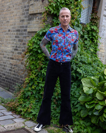 James is wearing Black Corduroy Bell Bottom Flares, paired with Hummingbird Print Short Sleeve Shirt and white trainers. He is posing with hands on his hips.
