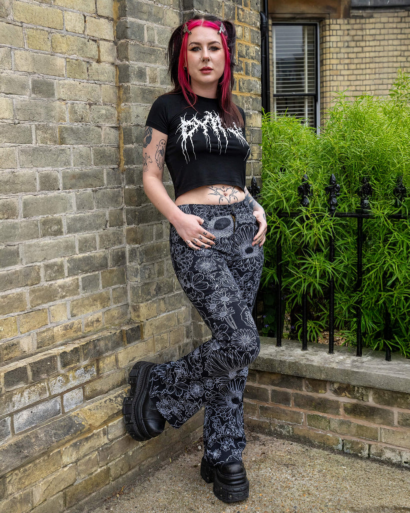 The Black and White Floral Bell Bottom Flares worn by a femme model with red and black hair wearing a black crop top and platform black boots. They are stood outside along a street in front of a black railing, a row of tall houses and green flowering bushes. They are facing right leaning back on the brick wall behind them with one leg bent. The black base flares has multiple floral white outlines all over.