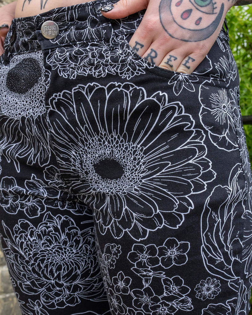 Close up of the Black and White Floral Bell Bottom Flares worn by a femme model with both hands resting in the front pockets highlighting the run & fly silver button detailing. The black base flares has multiple floral white outlines all over.