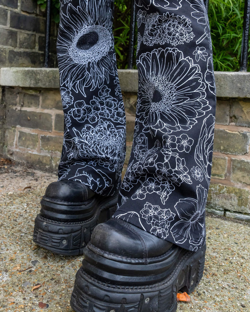 Close up of the Black and White Floral Bell Bottom Flares worn by a femme model with platform black boots highlighting the flared bell bottoms of the trousers. The black base flares has multiple floral white outlines all over.