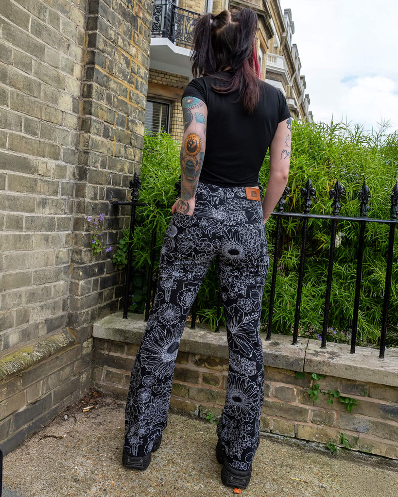 The Black and White Floral Bell Bottom Flares worn by a femme model with red and black hair wearing a black crop top and platform black boots. They are stood outside along a street in front of a black railing, a row of tall houses and green flowering bushes. They are facing away with one hand in the back pocket. The black base flares has multiple floral white outlines all over.