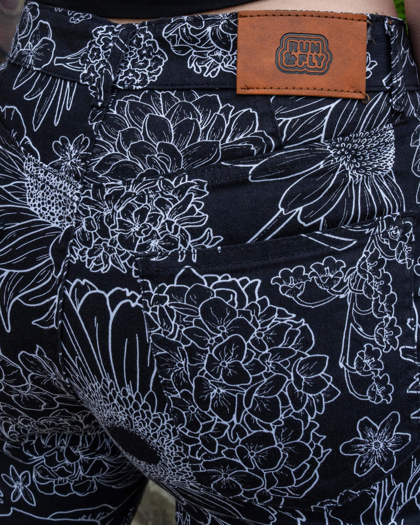 Close up of the Black and White Floral Bell Bottom Flares worn by a femme model facing away to highlight the run & fly faux leather brown tag on the back waistband. The black base flares has multiple floral white outlines all over.