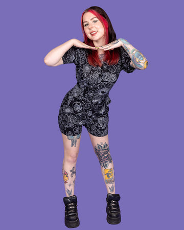The Black Floral Stretch Twill Playsuit worn by a femme model with mid length red and black hair with black platform trainers on a purple studio background. They are facing forward smiling leaning forward with their hands framing underneath their face. The black base playsuit has white floral outlines all over.