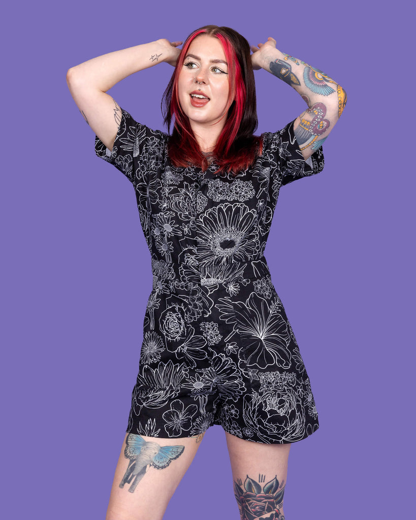 Close up of the Black Floral Stretch Twill Playsuit worn by a femme model with mid length red and black hair on a purple studio background. They are facing forward laughing looking up to the left with both hands resting on the back of her head. The black base playsuit has white floral outlines all over.